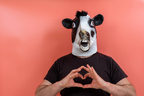 pessoa fantasiada usando uma máscara de vaca fazendo uma forma de coração com as mãos em um fundo rosa - Foto, Imagem