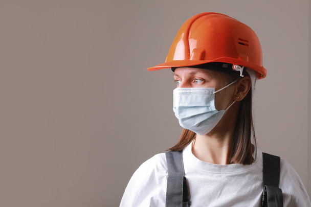 医療マスクとグレーの背景にオレンジの建設ヘルメットでかなりの女性労働者。コロナウイルスの顔マスク付き労働安全衛生の専門家｜covid-19. - 写真・画像
