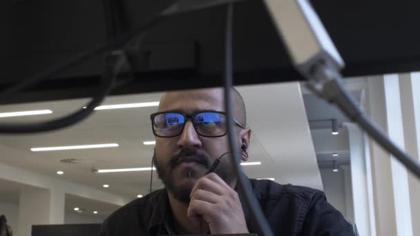 Minoría étnica Adulto Hombre con gafas en la oficina Hablando en el micrófono de auriculares. Visto desde detrás del monitor. Ángulo bajo, bloqueado - Metraje, vídeo