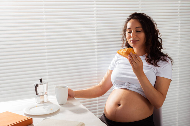 Радостная беременная молодая красивая женщина ест круассан и пьет чай во время утреннего завтрака. Концепция приятного утра и ожидания встречи с малышом. Copyspace - Фото, изображение