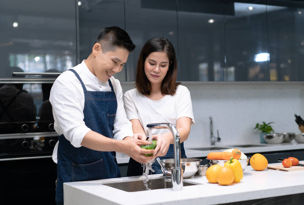 Portrait of young Asian couple in love while washing vegetables for cooking in kitchen at home. Couple spending time together at home. - Фото, изображение