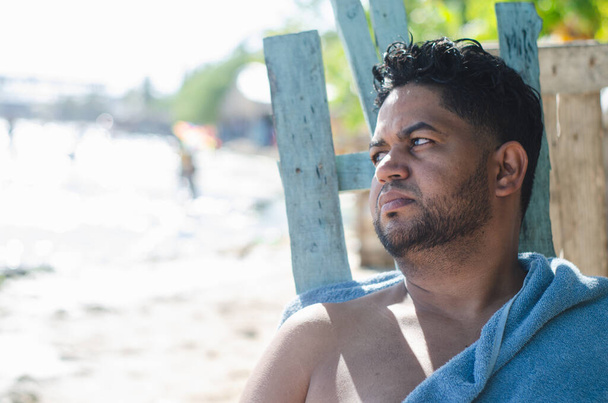 latino oscuro de la república dominicana descansando su día libre en la playa con una toalla y pensando - Foto, Imagen
