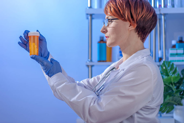 Ženský lékař se stylovými krátkými vlasy v bílém lékařském kabátě při pohledu na oranžový předpis sklenice pilulek na klinice. - Fotografie, Obrázek