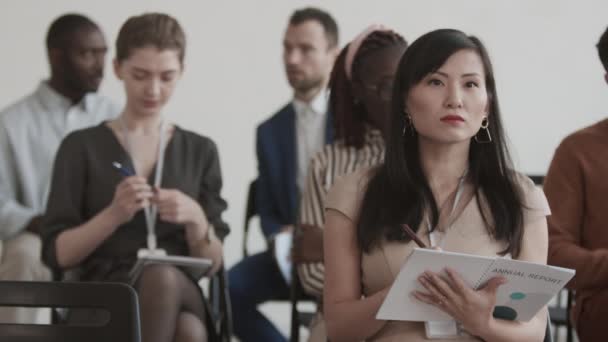 Gros plan moyen de la jeune femme d'affaires asiatique assise dans la salle de conférence au premier plan de divers collègues, prenant des notes dans le livre de rapport annuel - Séquence, vidéo