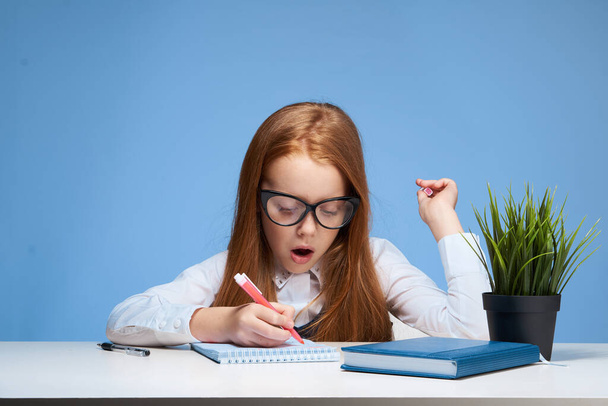 руда дівчина в окулярах робить домашнє завдання за столом шкільна освіта
 - Фото, зображення