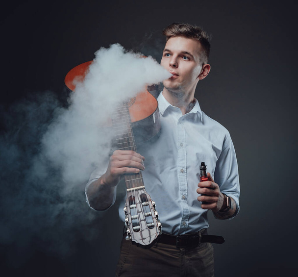 Modny facet z gitarą palenia papierosa elektrycznego w ciemnym tle - Zdjęcie, obraz