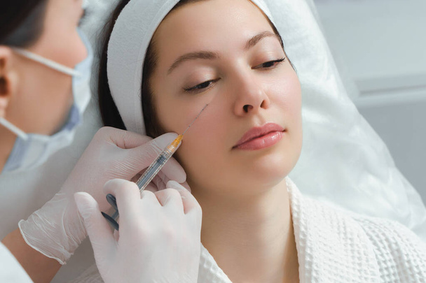 De arts cosmetoloog maakt de verjongende gezichtsinjecties procedure voor aanscherping en gladstrijken rimpels op de gezichtshuid van een mooie, jonge vrouw in een schoonheidssalon. Cosmetologie, huidverzorging - Foto, afbeelding