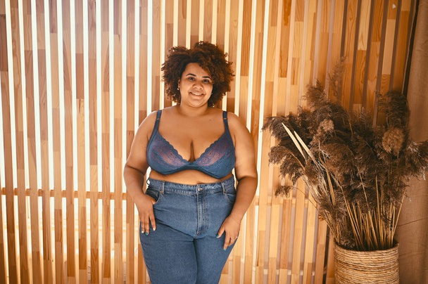 Beautiful stylish curvy oversize African black woman with afro hair posing in bra and jeans on wooden eco friendly background, body imperfection, body acceptance, body positive and diversity concept. - Photo, Image