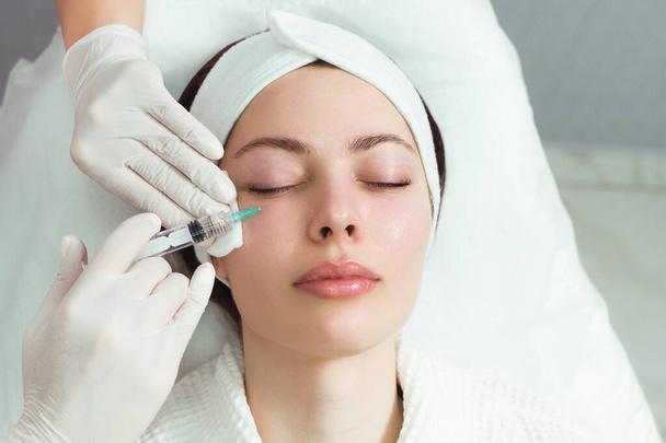 The doctor cosmetologist makes the rejuvenating facial injections procedure for tightening and smoothing wrinkles on the face skin of a beautiful, young woman in a beauty salon. Cosmetology, skincare - Photo, Image