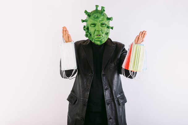 Persona disfrazada de coronavirus con una máscara de látex - virus covid-19, vistiendo una levita de cuero negro y una camiseta negra, sosteniendo máscaras quirúrgicas con ambas manos, sobre fondo blanco. Concepto de Coronavirus - Foto, imagen