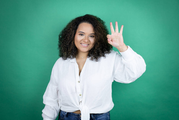 jong Afrikaans amerikaans meisje dragen wit shirt over groene achtergrond doen ok teken met vingers en glimlach, uitstekend symbool - Foto, afbeelding