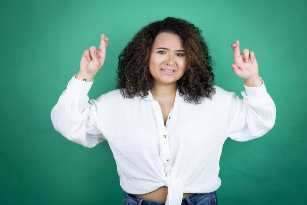 Jeune fille afro-américaine vêtue d'une chemise blanche sur fond vert geste doigt croisé souriant d'espoir et regardant côté - Photo, image