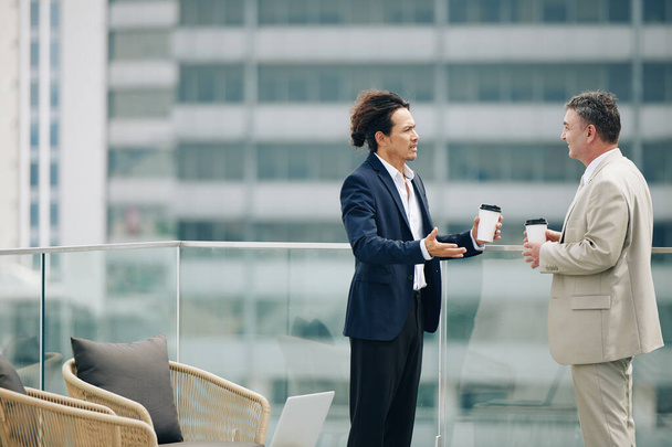 Хмурый бизнесмен смешанной расы с чашкой кофе разговаривает со зрелым коллегой, когда они встречаются на крыше - Фото, изображение