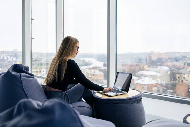 Красива молода жінка-менеджер сидить з ноутбуком на м'якому пуфі біля панорамного вікна. Дівчина бізнесмен працює над новим проектом
 - Фото, зображення