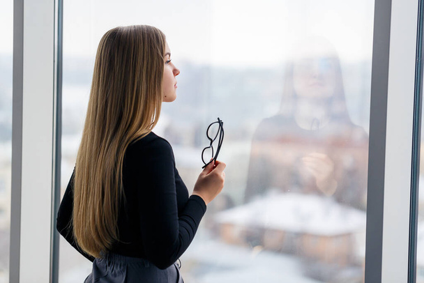 Μια επιτυχημένη γυναίκα μάνατζερ στο γραφείο της με μεγάλα παράθυρα στέκεται και κοιτάζει την πόλη. Επιχειρηματίας με γυαλιά στο παρασκήνιο των μεγάλων παραθύρων - Φωτογραφία, εικόνα