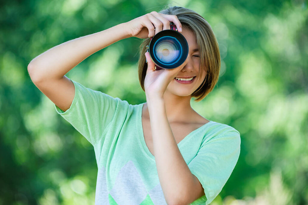 Νεαρή γοητευτική ήρεμη γυναίκα με πράσινη μπλούζα φωτογραφίζει μια κάμερα με ένα μεγάλο φακό στο καλοκαιρινό πάρκο. - Φωτογραφία, εικόνα