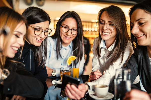 Side view of adult caucasian woman with eyeglasses holding mobile phone while sitting at cafe or restaurant with her female millennial friends making a video call on smartphone or taking selfie photo - Photo, Image