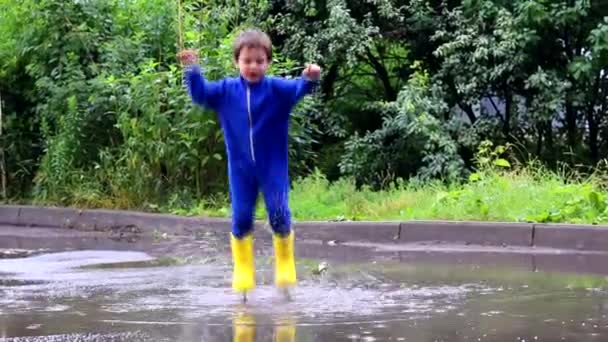 その少年は水たまりに飛び込む。ゴムのブーツを着た少年。幸せな子供時代。夏だ。夏の水たまり。雨の後に新鮮な。子供の楽しみ. - 映像、動画