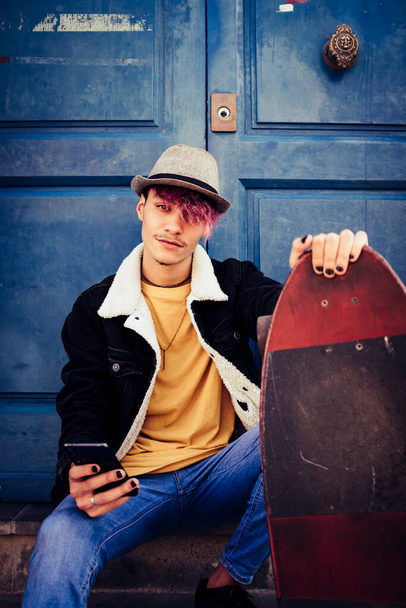 Diverso joven con pelo violeta y estilo de look alternativo utilizar el teléfono moderno sentado fuera con puertas azules en el fondo y skate board para moverse en la ciudad - Foto, imagen