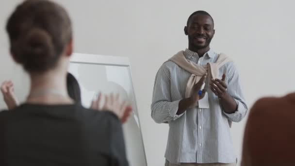Keskikokoinen olkansa yli nuori Afrikkalainen liikemies hymyilee, seisoo lähellä valkotaulu kuvaaja, taputtaa kädet tunnistamaton yleisö, istuu hänen edessään - Materiaali, video