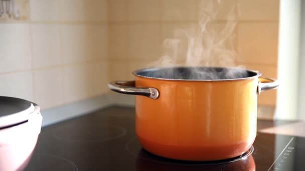 Closeup orange enamel steel cooking pan on modern inductive hob with boiling water or soup and scenic vapor steam backlit by warm sunlight at kitchen. Kitchenware utensil and tools at home background - Footage, Video