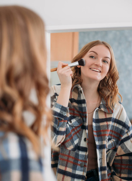 Femme caucasienne mignonne utilisant une brosse de maquillage et souriant devant le miroir sur fond de maison. Beauté, maquillage, cosmétique, concept matin et personnes - Photo, image