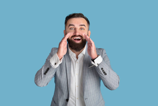 Возбужденный молодой офисный менеджер, кричащий руками у рта на фоне синей студии - Фото, изображение