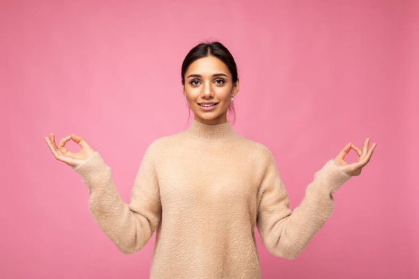 Jonge positieve kalme gelukkige aantrekkelijke brunette vrouw met oprechte emoties dragen beige trui geïsoleerd over roze achtergrond met kopieerruimte en het doen van bemiddeling. Yoga-concept - Foto, afbeelding