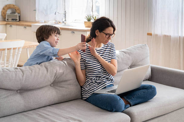Freelancer mamá trabajando en el ordenador portátil en casa durante el encierro, niño ruidoso distrae pidiendo atención. - Foto, imagen