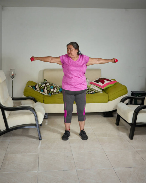 Kypsä nainen nostaa käsipainoja kotona. Vanhempi nainen seisoo tekemässä käsi venyttely käyttää kevyitä painoja.                                         - Valokuva, kuva
