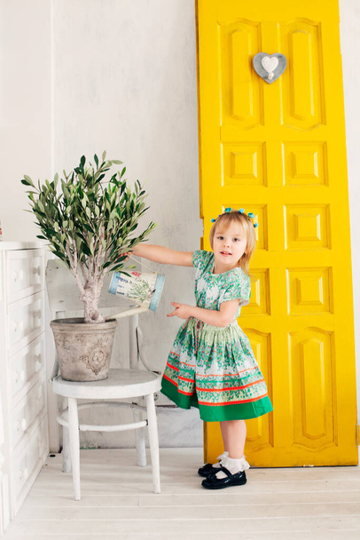 Beautiful little blonde girl in floral wreath on white and yellow kitchen background cares for, watering huge plant. Easter concept. Easter card. Color of the year - Photo, Image