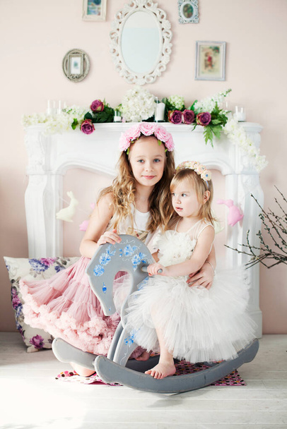 Dvě krásné dívky v květinových věncích a nádherné sukně sedí u krbu. Velikonoce, jarní koncepce - Fotografie, Obrázek