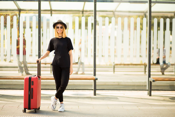 Retrato de comprimento total de viagem elegante jovem mulher desgaste em preto total com mala vermelha esperar transporte - Foto, Imagem