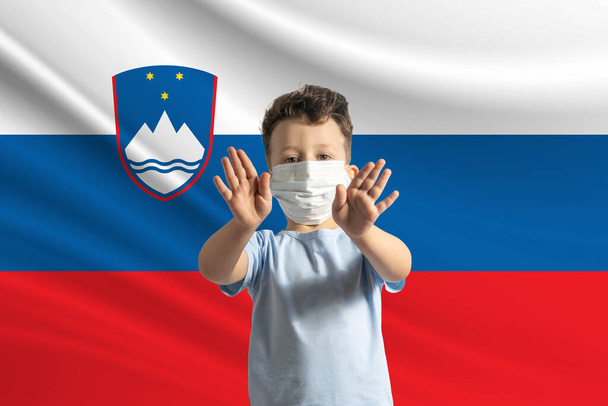 Μικρό λευκό αγόρι με προστατευτική μάσκα στο φόντο της σημαίας της Σλοβενίας. Κάνει στοπ με τα χέρια του, μένει στο σπίτι Σλοβενία. - Φωτογραφία, εικόνα