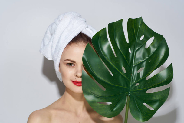 Una mujer con una toalla en la cabeza sostiene una hoja de palma cerca del maquillaje de la cara hombros desnudos cosmetología aspecto natural - Foto, imagen
