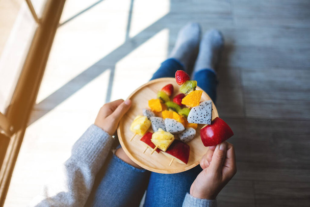 Vista superior de la imagen de una mujer comiendo y sosteniendo un plato de madera de frutas frescas mezcladas en pinchos mientras está sentada en el suelo - Foto, Imagen