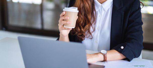 Closeup image of businesswoman using and typing on laptop while working and drinking coffee in office - Foto, afbeelding