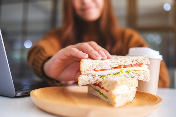Μια Ασιάτισσα που κρατάει και τρώει σάντουιτς ολικής άλεσης ενώ δουλεύει σε φορητό υπολογιστή - Φωτογραφία, εικόνα