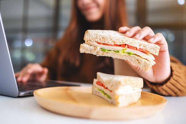 Азиатская женщина держит и ест сэндвич с пшеницей во время работы на ноутбуке - Фото, изображение