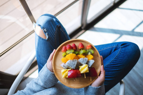 Imagen de vista superior de una mujer sosteniendo un plato de madera de frutas frescas mezcladas en pinchos - Foto, Imagen