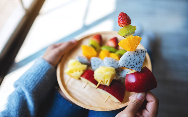 Closeup image of a woman holding a wooden plate of fresh mixed fruits on skewers - Photo, Image