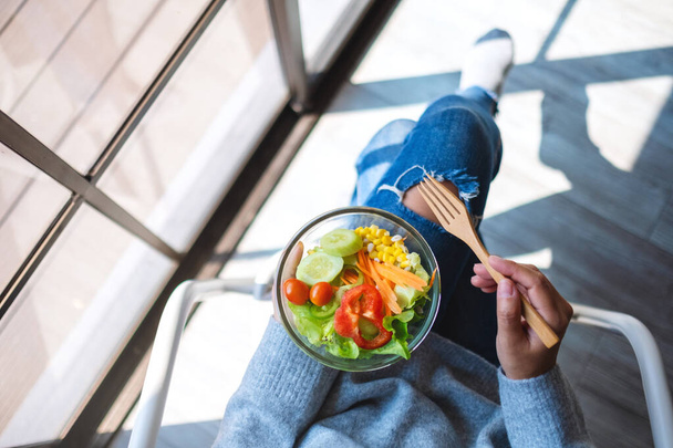 Bovenaanzicht beeld van een vrouw die een kom verse gemengde groenten salade vasthoudt terwijl ze op de stoel zit - Foto, afbeelding