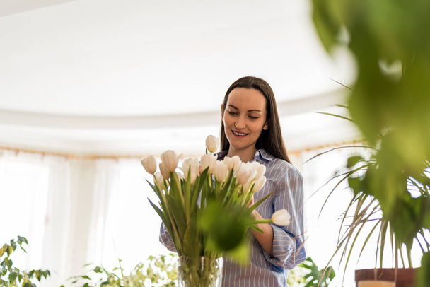 lächelnde glückliche Frau im blauen Hemd kümmert sich um weiße Tulpen, genießt das Leben, verbindet sich mit der Natur - Foto, Bild