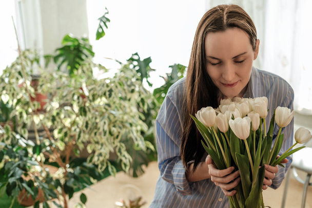 žena v modré košili očichává bílé tulipány, květinový skleník doma, koncept péče o květiny - Fotografie, Obrázek