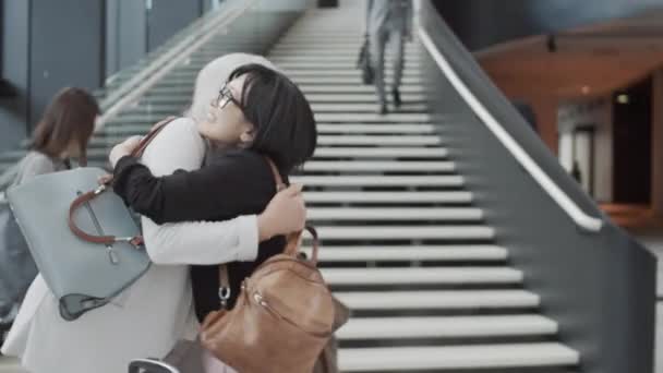 Kamerafahrt von glücklicher asiatischer Frau, die eine Freundin umarmt, die aus dem Ankunftstor des Flughafens geht, sowohl verlässt als auch plaudert, und anderen Passagieren mit herumrauschendem Gepäck - Filmmaterial, Video