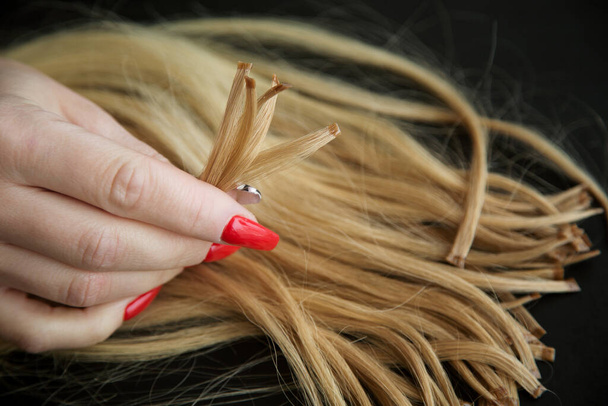 ingekapselde strengen blond haar in de handen van vrouwen met een felrode manicure. Het concept van haarverzorging en uitbreiding.  - Foto, afbeelding