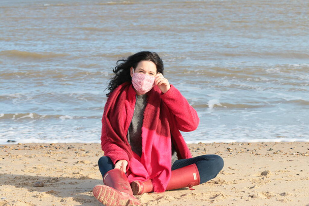 Attractive female wearing mask with black hair in red jacket scarf and brown leather boots seated cross legged on beautiful sandy beach by ocean in fresh air at Winterton On Sea Norfolk east Anglia UK in lockdown for Covid-19 virus sea background - Photo, Image