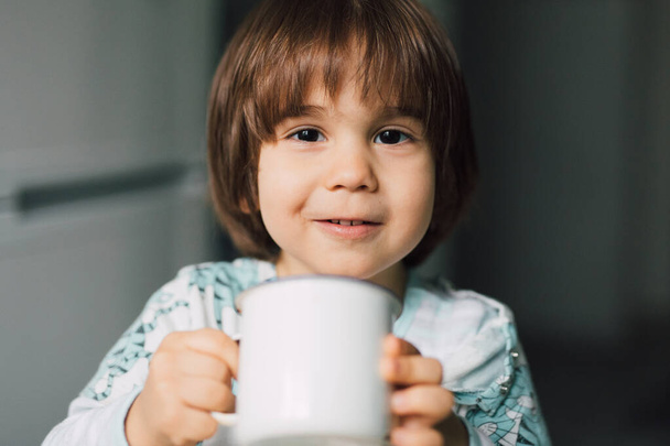 Evde elinde sütle kupa tutan sevimli, gülümseyen anaokulu çocuğu. Otantik gülüşlü çocuk, kahvaltı yapıyor.. - Fotoğraf, Görsel