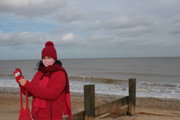Close up of female wearing mask with black hair and red clothes of red jacket scarf and hat seated on sandy beach by ocean in Winterton Norfolk east Anglia UK in lockdown for Covid-19 virus outbreak sea background - Photo, Image