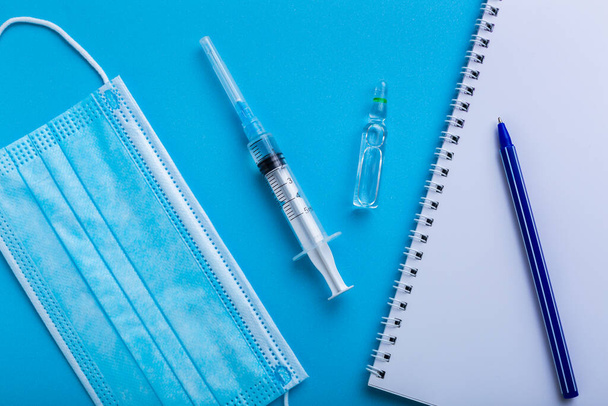 Een spuit met vaccin op blauwe achtergrond met een beschermend gezichtsmasker en witte zwarte noot voor een recept. - Foto, afbeelding